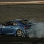 A blue sports car performing a drift maneuver on an open race track surrounded by smoke.